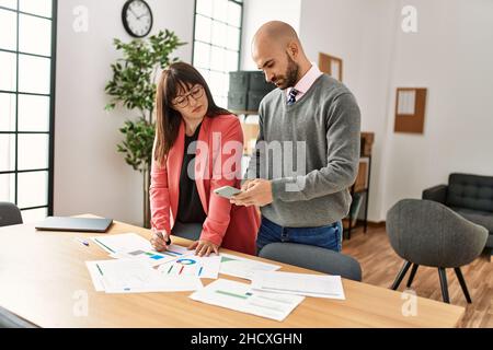 Deux professionnels hispaniques avec une expression sérieuse travaillant avec un smartphone au bureau. Banque D'Images
