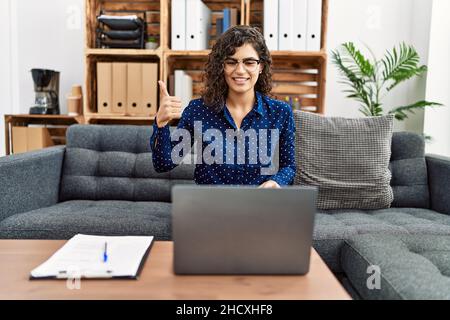Jeune femme brunette travaillant à la clinique de psychologie faisant un rendez-vous en ligne souriant heureux et positif, pouce vers le haut faisant excellent et signe d'approbation Banque D'Images