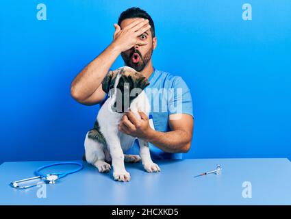 Beau vétérinaire hispanique avec la barbe contrôle de santé chien peeking dans le choc couvrant le visage et les yeux avec la main, regardant à travers les doigts avec l'embarr Banque D'Images