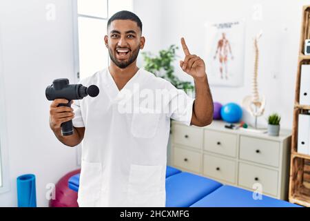 Un jeune kinésithérapeute indien tenant un pistolet de massage thérapeutique au centre de bien-être souriant émerveillé et surpris et pointant vers le haut avec les doigts et les bras relevés. Banque D'Images