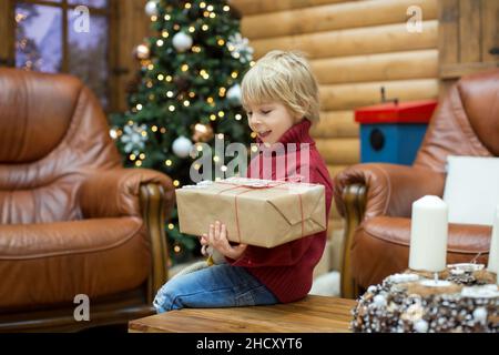 Enfant blond, livre de lecture et cadeaux de tenue dans un chalet confortable dans les montagnes, l'heure de Noël Banque D'Images