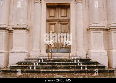 Des bouquets de fleurs se trouvent sur des plinthes dorées sur les marches humides devant une porte en bois de l'église Banque D'Images