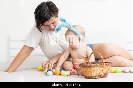 Maman et bébé avec des oreilles de lapin, avec des œufs de Pâques dans leurs mains, assis sur le canapé, parents et enfants jouent à l'intérieur. La famille fête Pâques Banque D'Images