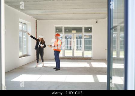 Femme ingénieur de bâtiment décrivant le travail sur la fenêtre de la salle à un ouvrier de construction, qui écrit dans le bloc-notes Banque D'Images