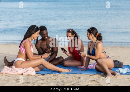 Joyeux homme multiethnique et des femmes souriant et parcourant les médias sociaux sur les téléphones mobiles tout en étant assis sur une couverture pendant un pique-nique sur un bord de mer sablonneux sur Lanzaro Banque D'Images