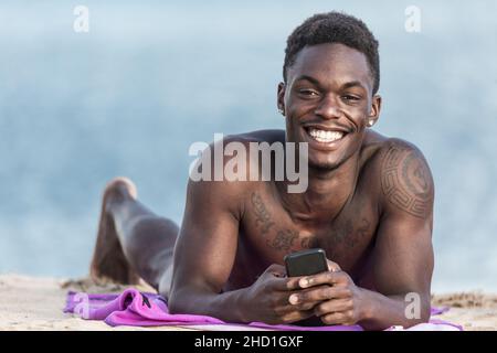 Joyeux Afro-américain tatoué homme avec le téléphone portable souriant et regardant la caméra tout en se relaxant sur la plage de sable sur fond flou de la mer sur Lanzar Banque D'Images