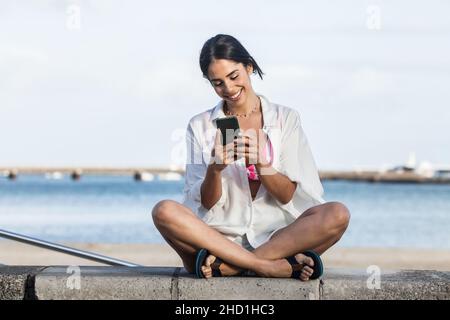 Femme souriante en vêtements d'été assise avec des jambes croisées sur la bordure en pierre et messagerie sur smartphone tout en appréciant le week-end à Lanzarote Banque D'Images