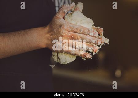 Un homme dans un tablier bleu tient la pâte de blé frais pour cuire dans ses mains.Le processus de préparation de la pâte.Cuisine maison. Banque D'Images