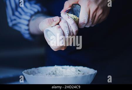 Un cuisinier dans un tablier bleu et une chemise rayée tient un moulin à poivre dans ses mains, et assaisonner la farine pour la pâte avec elle.Le processus de la cuisine à domicile. Banque D'Images
