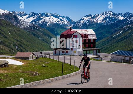 Un cycliste qui monte sur la route du col de Furka, en passant par l'hôtel Galenstock, des montagnes enneigées au-dessus d'Andermatt au loin. Banque D'Images
