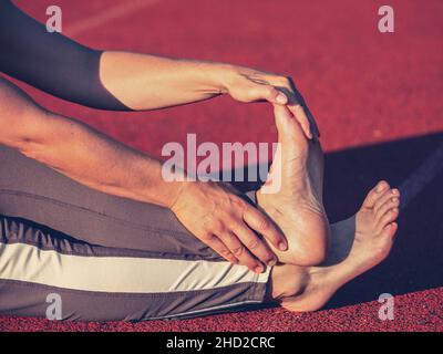 Femme adulte pieds nus étirant les tendons et les chevilles sur la piste de course rouge au soleil du soir.Corps d'âge moyen avec longs leggings Banque D'Images