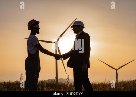 Silhouette d'un ingénieur afro-américain et d'un inspecteur indien utilisant une tablette numérique lors d'une réunion à l'extérieur et en serrant la main.Deux partenaires debout sur le terrain avec des éoliennes pendant un coucher de soleil étonnant. Banque D'Images