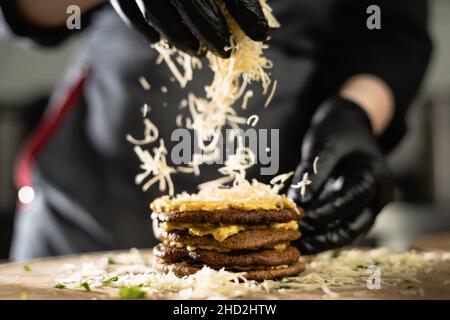 le chef prépare une tarte au foie dans la cuisine.Gros plan des mains de la femme en gants parsemer de crêpes au foie de fromage. Banque D'Images