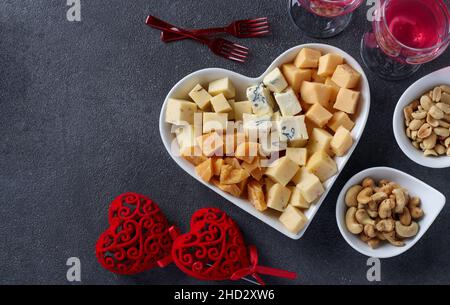 Variétés de fromages d'élite dans une assiette en forme de cœur, noix de cajou, arachides et deux verres de vin pour la Saint-Valentin sur fond gris, vue du dessus Banque D'Images