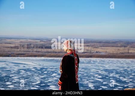 Vue arrière de la femme anonyme en robe traditionnelle et harnais touchant le visage et en admirant la rivière gelée contre le ciel bleu le froid hiver dans la nature Banque D'Images