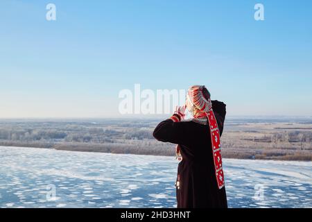 Vue arrière de la femme anonyme en robe traditionnelle et harnais touchant le visage et en admirant la rivière gelée contre le ciel bleu le froid hiver dans la nature Banque D'Images
