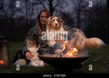 Une jeune femme est assise à l'extérieur dans les bois avec ses deux chiens de berger australiens.Neige sur l'herbe, la nuit près du feu de camp.Elle embrasse les chiens Banque D'Images
