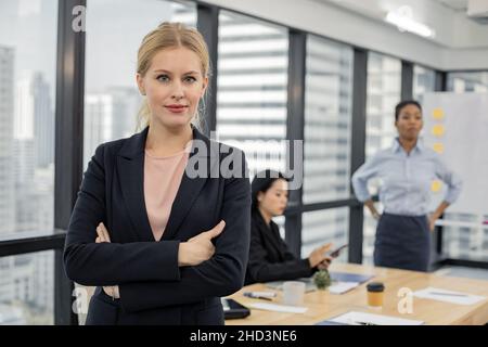 Belle femme au bureau avec les bras pliés et l'expression confiante que les autres travailleurs tiennent une réunion en arrière-plan.Portrait d'une femme d'affaires caucasienne Banque D'Images