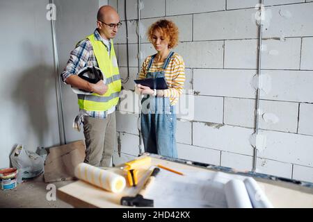 S'est concentré sur l'équipe d'ingénierie des tâches qui se penche sur un plan, en discutant.À côté d'un mur de briques à l'intérieur d'un bâtiment en construction. Banque D'Images