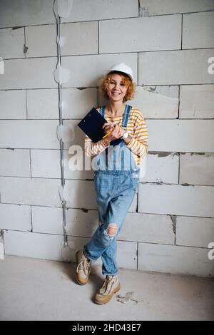 Femme rayonnante souriante ingénieur de construction dans un casque blanc tenant un presse-papiers, penchée sur un mur de briques gris rugueux. Banque D'Images