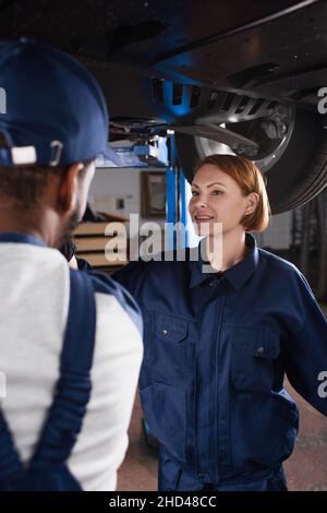Un mécanicien souriant en uniforme parlant avec un collègue afro-américain flou sous la voiture dans un garage Banque D'Images