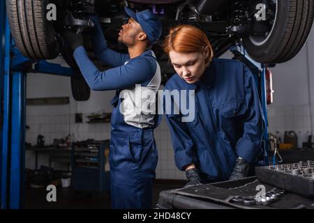 Mécanicien debout près des outils et collègue afro-américain travaillant avec la voiture dans le garage Banque D'Images