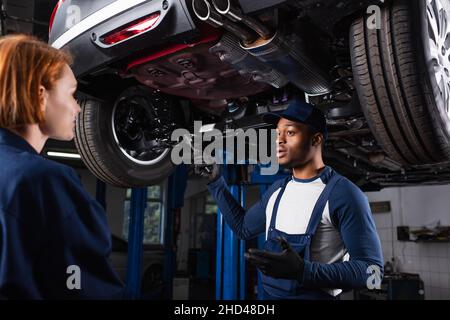 Mécanicien afro-américain tenant la clé en parlant à un collègue sous la voiture dans un garage Banque D'Images