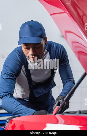 jeune technicien afro-américain en combinaison et casquette inspectant la voiture avec capot ouvert Banque D'Images