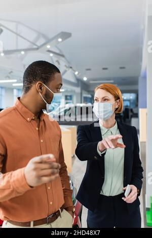 client afro-américain et concessionnaire automobile dans les masques médicaux se regardant dans la salle d'exposition Banque D'Images