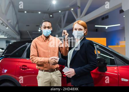 concessionnaire automobile et client afro-américain dans les masques médicaux qui se secouent les mains tout en regardant la caméra Banque D'Images