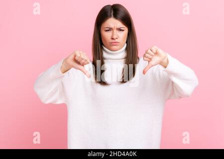 Portrait de la triste brune femelle montrant le pouce vers le bas démontrant le geste de désapprobation et de dégoût, portant blanc style décontracté pull.Studio d'intérieur isolé sur fond rose. Banque D'Images