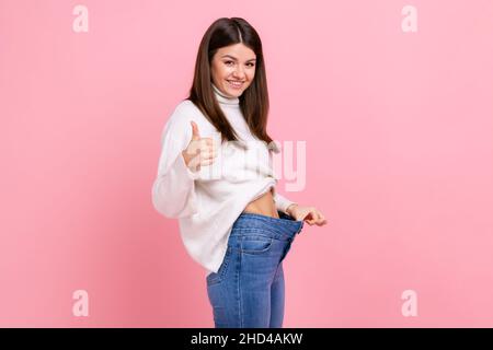 Portrait de côté de la belle femme heureuse montrant le pouce vers le haut et sa taille mince et grand pantalon, portant blanc style décontracté pull.Studio d'intérieur isolé sur fond rose. Banque D'Images