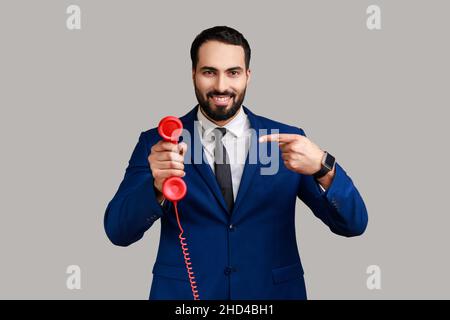 Homme barbu pointant le doigt sur le combiné d'un téléphone fixe rouge vintage, intéressé par des appareils rétro, répondant aux appels, portant un costume de style officiel.Prise de vue en studio isolée sur fond gris. Banque D'Images