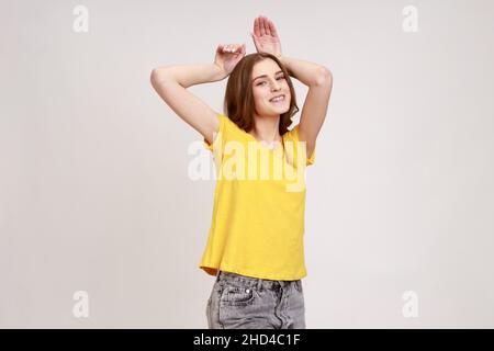 Portrait de drôle insouciant belle adolescente fille dans un T-shirt jaune montrant le geste des oreilles de lapin et regardant ludique heureux, comportement puéril.Prise de vue en studio isolée sur fond gris. Banque D'Images