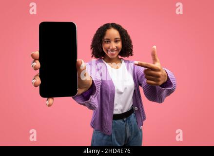 Joyeuse femme noire pointant vers le téléphone portable avec écran vide, publicité nouvelle application ou site Web, offrant maquette pour votre annonce Banque D'Images