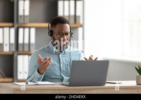 Mécontent choqué choqué jeune afro-américain barbu des lunettes, des gestes de casque et des travaux sur pc Banque D'Images