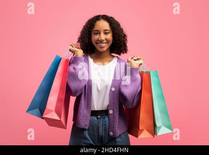 Charmante jeune femme noire avec des sacs de shopping souriant à l'appareil photo sur fond rose studio Banque D'Images