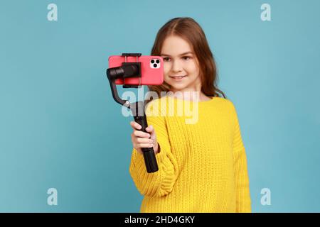 Portrait de petite fille avec expression positive, tenant Steadicam et smartphone, streaming, portant jaune style décontracté pull.Studio d'intérieur isolé sur fond bleu. Banque D'Images