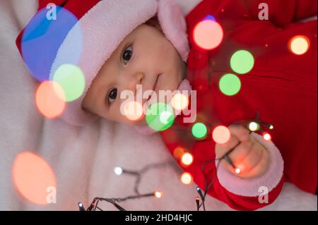 Un petit garçon habillé d'un costume de père noël se trouve sur le lit et joue avec une guirlande de Noël. Banque D'Images