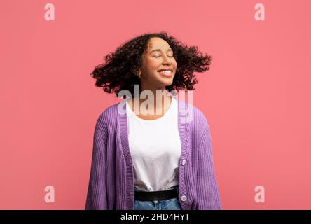 Portrait d'une femme afro-américaine souriante posant avec des cheveux volants et sains sur fond rose studio Banque D'Images