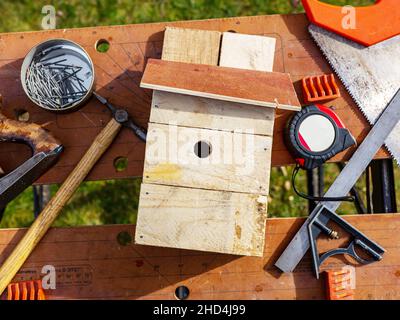 Une boîte à oiseaux faite à la main, posée sur un établi avec les outils utilisés qui l'entourent, comprenant un marteau et des clous, une fraiseuse à main, une scie à main et un mètre à ruban Banque D'Images