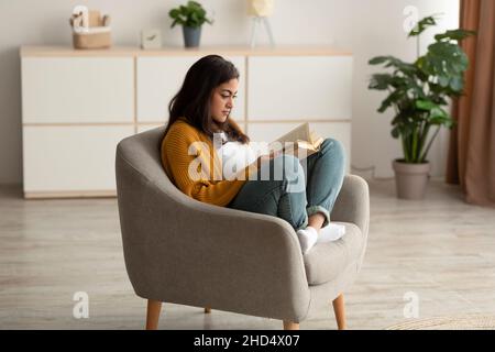 Femme arabe concentrée assise dans un fauteuil, lisant un livre dans la salle de séjour, appréciant une matinée tranquille avec une histoire intéressante Banque D'Images