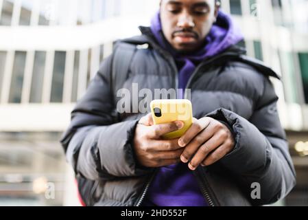 Attention douce de l'homme afro-américain dans les vêtements d'extérieur de navigation de téléphone cellulaire avec boîtier jaune tout en se tenant dans la rue de la ville moderne Banque D'Images
