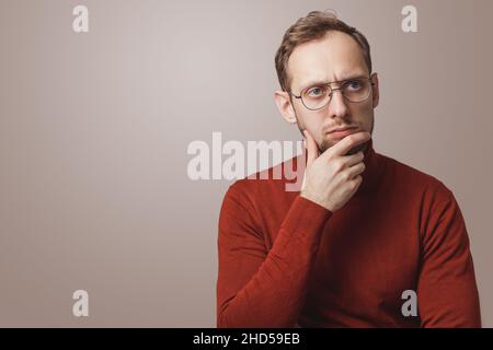 Un jeune homme au jeune âge tient le menton et regarde pensivement loin porte des lunettes et un col roulé rouge sur fond gris Banque D'Images
