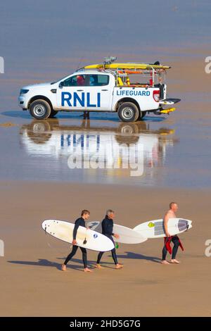 Surfeurs transportant leurs planches de surf et passant devant un véhicule d'intervention d'urgence RNLI sur la plage de Fistral à Newquay, en Cornouailles. Banque D'Images