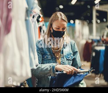 Femme dans un masque médical choisissant des vêtements dans un magasin d'usine. Banque D'Images