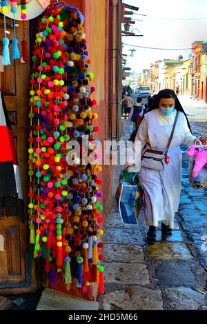 Façade avec des marchandises colorées accrochées le long de la porte ouverte de la rue et trottoir avec une nonne dans son habitude avec piñata à San Miguel de Allende, MX Banque D'Images