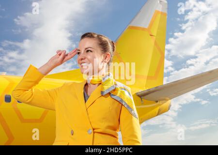 Un accompagnateur de vol souriant en uniforme de compagnie aérienne fait un geste de salut et souriant tout en se tenant près d'un avion jaune Banque D'Images