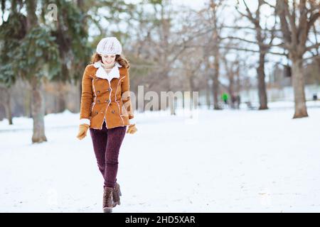 Portrait complet d'une femme moderne souriante de 40 ans avec des moufles dans un bonnet tricoté et un manteau de peau de mouton marchant à l'extérieur dans le parc de la ville en hiver Banque D'Images