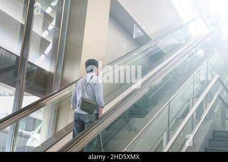 Homme asiatique voyageur avec masque facial sur l'escalier roulant dans un bâtiment moderne Banque D'Images